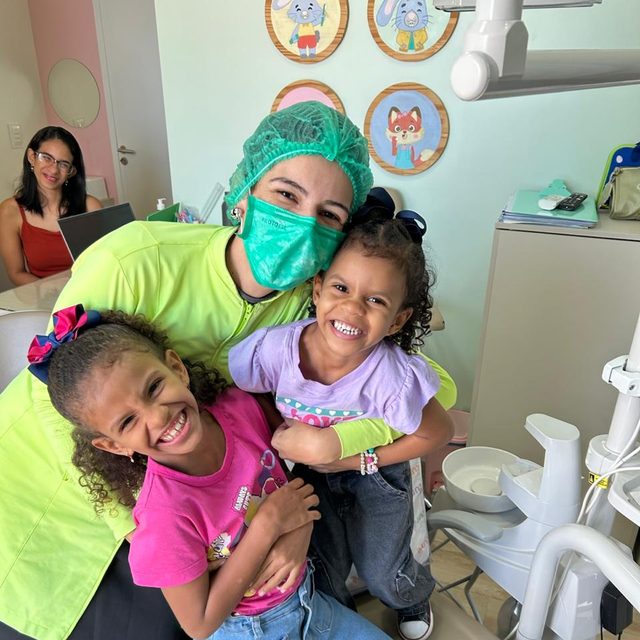 Duas irmãs abraçadas com a dentista, rindo na cadeira