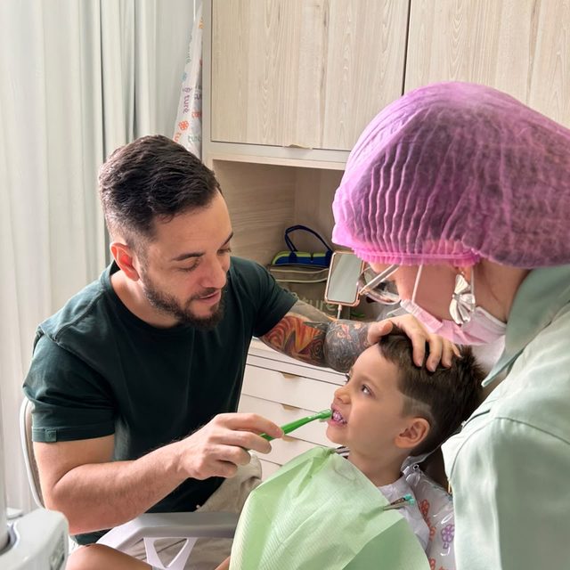 Pai escovando os dentes do filho enquanto a dentista segura a cabeça dele